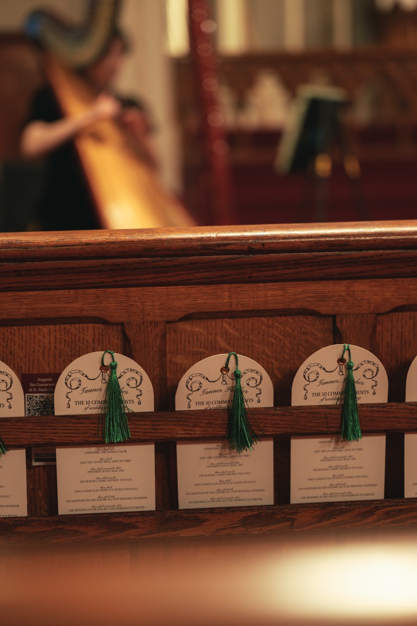 Custom ceremony programs with green tassels placed in church pews for Tanner Fletcher's bridal fashion show at New York Bridal Fashion Week, with a harpist performing in the background.